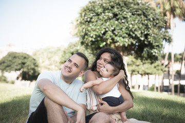 Fototapeta premium Mother, father and baby sitting on the grass of the public park. Happy young family spending time together outside - Image