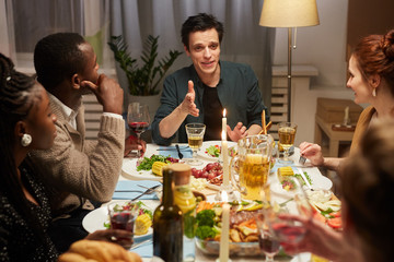 Group of friends sitting at the table and listening to young man who talking during holiday dinner