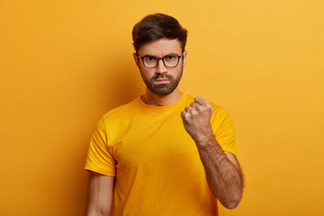 Strict bearded man looks seriously at camera, shows fist with angry expression, wears casual yellow t shirt and spectacles, isolated on yellow background, makes furious grimace, stands displeased