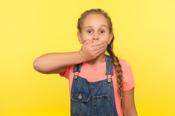 I won't tell! Portrait of scared little girl with braid in denim overalls covering mouth with hand, afraid to say secret, child terrified to speak. indoor studio shot isolated on yellow background
