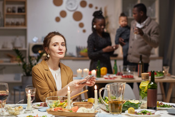 Portrait of young woman looking at camera while sitting at holiday table and using her mobile phone with her friends in the background