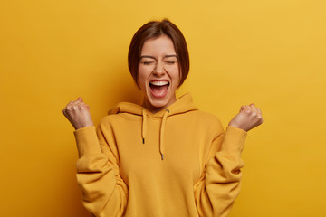 Lucky positive woman feels happy and relieved, achieves reward, clenches fists with triumph, celebrates great amazing news, exclaims with joy, wears casual hoodie, isolated on yellow background