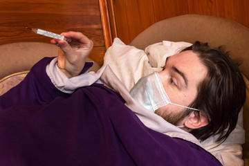 a man with dark hair and a disposable mask to protect against the virus, lies at home in bed, isolating himself from other people. fever, headache and nausea are symptoms - take care of your health