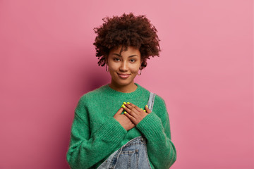 Touched thankful ethnic woman keeps hands on heart, expresses gratitude and kindness for nice gift, smiles gently, wears green sweater and overalls, thanks for help, poses against pink background. - obrazy, fototapety, plakaty