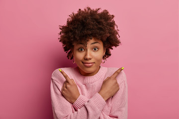 Indecisive pleased woman with curly hair, crosses hands over chest, points sideways unsure, has questioned expression and asks advice, wears casual pink jumper, poses indoor, faces decision.