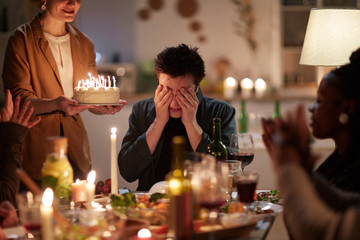 Young man sitting at the table and closing his eyes with hands while his friends carrying birthday cake with candles