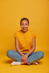 Fototapeta na wymiar Full length shot of cheerful happy dark skinned ethnic woman sits with legs crossed braided hair has positive smile on face, wears casual t shirt, jeans and socks, isolated on yellow studio background