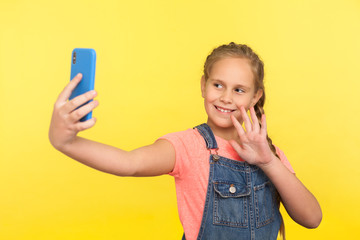 Hello! Portrait of adorable little girl in denim overalls gesturing hi while communicating with parents on video call, taking selfie on mobile phone. indoor studio shot isolated on yellow background