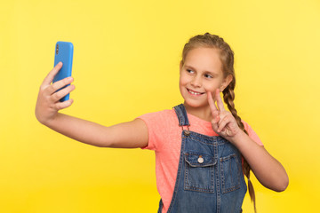 Portrait of adorable little girl in denim overalls gesturing victory or peace while communicating with parents on video call, taking selfie on mobile phone. studio shot isolated on yellow background