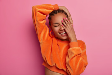 Portrait of happy dark skinned woman covers face with palm, keeps eyes shut, smiles tenderly, wears orange top, isolated on pink background, expresses happiness. Positive emotions and feeilngs - obrazy, fototapety, plakaty