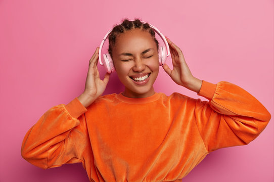 Gorgeous cheerful ethnic female smiles happily, listens music with headphones, closes eyes, enjoys loud sound, has cornnrow braided hairstyle, wears velvet bright orange sweater, poses indoor