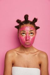 Close up shot of sad Afro American woman gets facial treatment, has clean fresh skin, looks upwards with unhappy expression, stands wrapped in towel, isolated on pink background. Beauty concept