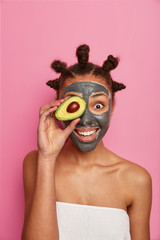 Taking care of your skin in natural way. Positive young woman applies clay mask on face, holds piece of avocado in front of eye, smiles happily, enjoys cosmetic treatment, cares about complexion