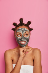 Pleasant looking ethnic woman touches chin gently, smiles positively and looks aside, applies face mask, awaits for nice result after beauty procedures, poses wrapped in towel, isolated on pink wall