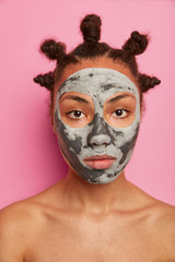 Close up shot of serious dark skinned woman applies beauty face mask for reducing pores and blackheads, cares about complexion and body, stands topless, looks directly at camera, has many hair buns