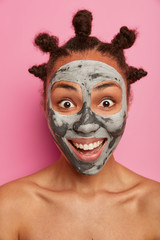 Close up portrait of shirtless positive woman has happy surprised reaction, beaming smile, perfect white teeth, wears clay mask for smooth face skin, isolated on pink background. Wellness concept