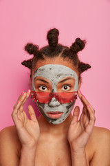 Image of dark skinned feminine girl applies face mask, looks surprisingly through pink sunglasses, gets beauty or cosmetic treatments, has bare shoulders, poses indoor, enjoys effective good result