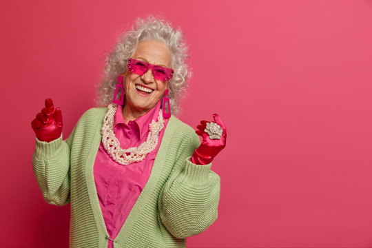 Joyful curly elderly woman dances carefree, has fun, wears trendy sunglasses, dressed in fashionable clothes, enjoys life, models against pink background, copy space for your advertisement or text