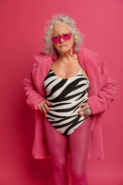 Vertical image of strict elderly woman in sunglasses keeps hands on waist, wears swimsuit, coat and pink tights, looks seriously at camera, waits for explanations, isolated on pink background