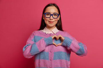 Pleasant looking tender woman confesses in love, makes heart sign, likes someone, expresses good feelings, wears transparent spectacles and striped jumper, shows sympathy, has romantic relationship