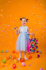 happy little girl in a blue dress holds a gift bag and catches confetti while standing on a yellow background. holiday concept, space for text