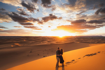 Fototapeta na wymiar Couple in love kissing in Sahara desert dunes, Morocco. Happiness, freedom and escape concept. Amazing warm sunset with beautiful colorful clouds.