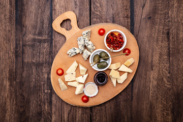 Different types of delicious cheese, olives, jam and sun-dried tomatoes on a wooden board. Top view.