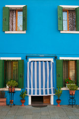 Bright blue facade of a house on the island of Burano in Italy. Entrance to a blue house with green shutters.