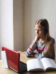 A blonde girl with headphones sits at a table with a computer and a book. Home schooling. The school is closed on quarantine due to the pandemic of Covid19. Distance learning