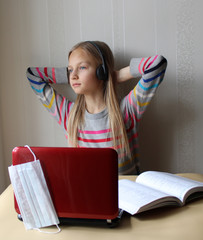 A blonde girl with headphones sits at a table with a computer and a book. Home schooling. The school is closed on quarantine due to the pandemic of Covid19. she's bored. Distance learning