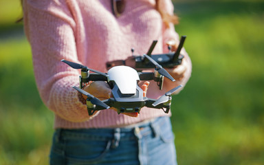 A young woman in a clearing in a park launches, checks, holds a drone, in one hand, in the other control panel.