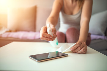 Fototapeta premium Cropped shot of an unrecognizable woman cleaning her device screen at home. Young person cleaning a mobile phone display by spraying washing solution. Woman holding a smartphone