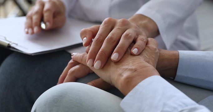 Close up young nurse holding hand of elderly patient, supporting after bad diagnosis at consultation. Empathic social worker in uniform comforting mature old woman, giving psychological help.