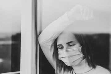 Coronavirus. Sick woman wearing protection face mask looking through the window. Patient isolated in house to prevent infection. Quarantine room at hospital. Black and white photo.