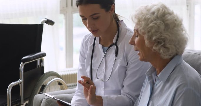 Focused young physiotherapist consulting mature disabled woman. Smiling doctor filling handicapped patient card form at checkup at home, listening to complaints, helping with rehabilitation process.
