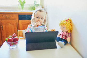 Adorable toddler girl eating fresh strawberries and using tablet. Child watching cartoons while eating. Digital technologies in kids education