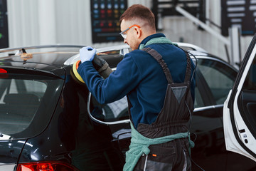 Male worker in uniform polishing new modern car. Conception of service