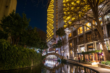 Naklejka premium The popular riverwalk in a night scene in downtown San Antonio, Texas, with an arch bridge and tall buildings along the river reflected in the water.