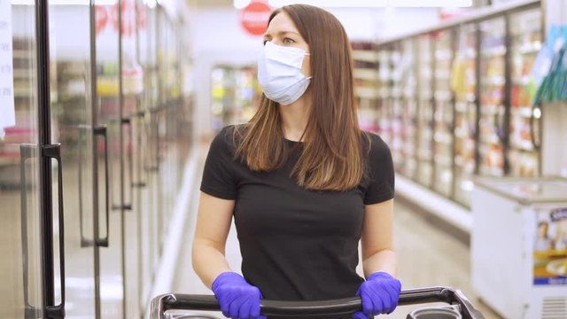 Woman shopping in grocery store for food while wearing PPE and preventing spread of virus germs by wearing face mask and latex gloves