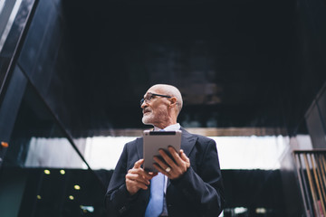 Intelligent senior businessman in suit and eyeglasses browsing tablet in front of business center on street
