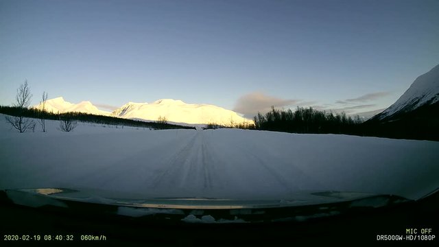 view from a dashcam on a snow-covered white ice road in northern norway with a shaded landscape and sunny mountains in the background