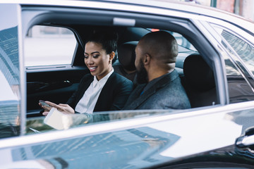 Satisfied executive lady using phone discussing project with black coworker in car