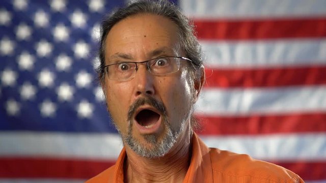 Tight portrait of mature man with beard wearing an orange shirt and looking shocked and surprised with out of focus American flag in the background.
