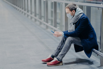 Tired tourist takes break, uses mobile phone, dressed in jacket, jeans and sneakers, wears protective mask against coronavirus, cares about health in public place, reads information. Safety concept