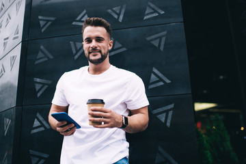 Confident man using smartphone and holding cup of coffee while looking at camera