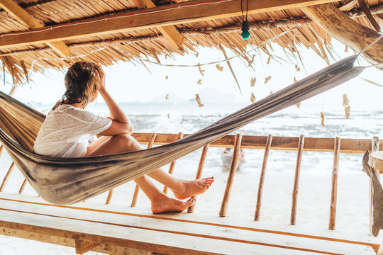 Young female sitting in hammock on straw hut terrace and enjoying a seaside beach landscape on Tonsai Beach in Thailand. Careless summertime exotic vacation concept image.