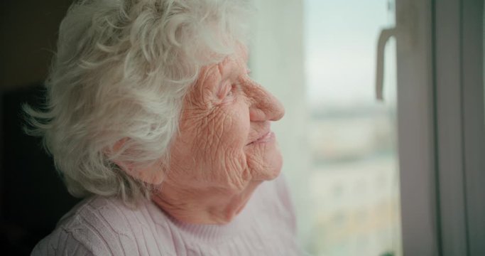 Old woman with deep wrinkles. Grandmother with white hair looks out the window and smiles.