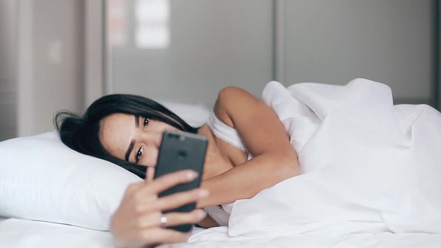 Pretty young girl in a bed takes selfies using a smartphone