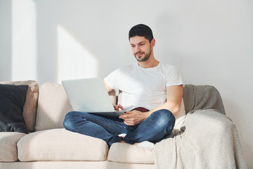 Man in white shirt and jeans sitting on bed with laptop indoors