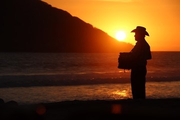 Hombre mexicano con acordeón a contraluz durante el atarceder en la playa, Mazatlán, Sinaloa,...